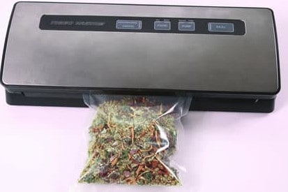 Vacuum sealer machine sealing a plastic bag filled with dried herbs and vegetables, with the bag partially inserted into the device.