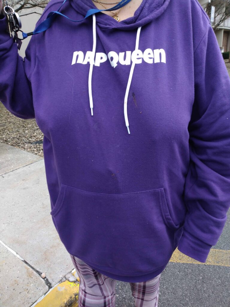 Woman wearing a purple hoodie with white drawstrings and text on the front, standing outdoors on a sidewalk, holding keys, with patterned leggings visible.