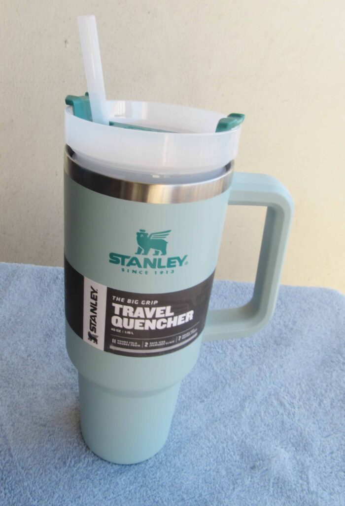 Light blue Stanley travel tumbler with handle and straw, labeled Big Grip Travel Quencher, sitting on a soft blue towel.