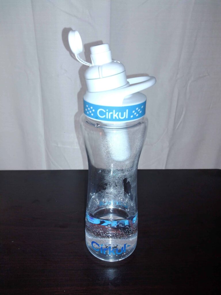 Clear Cirkul water bottle with white lid and flip-top cap, partially filled with water and condensation, placed on a dark surface.