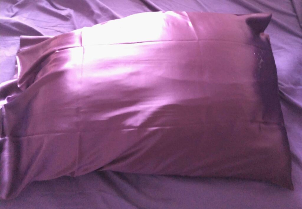 Purple satin pillowcase on a bed, showing a smooth, shiny surface with soft light reflecting across the fabric.