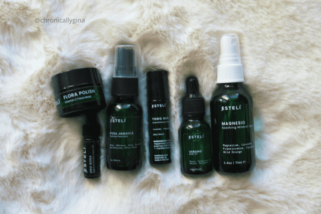 Esteli Skin Care products including face mask, facial oils, and sprays arranged on a soft beige fur background, showcasing the full collection in dark green bottles and jars.