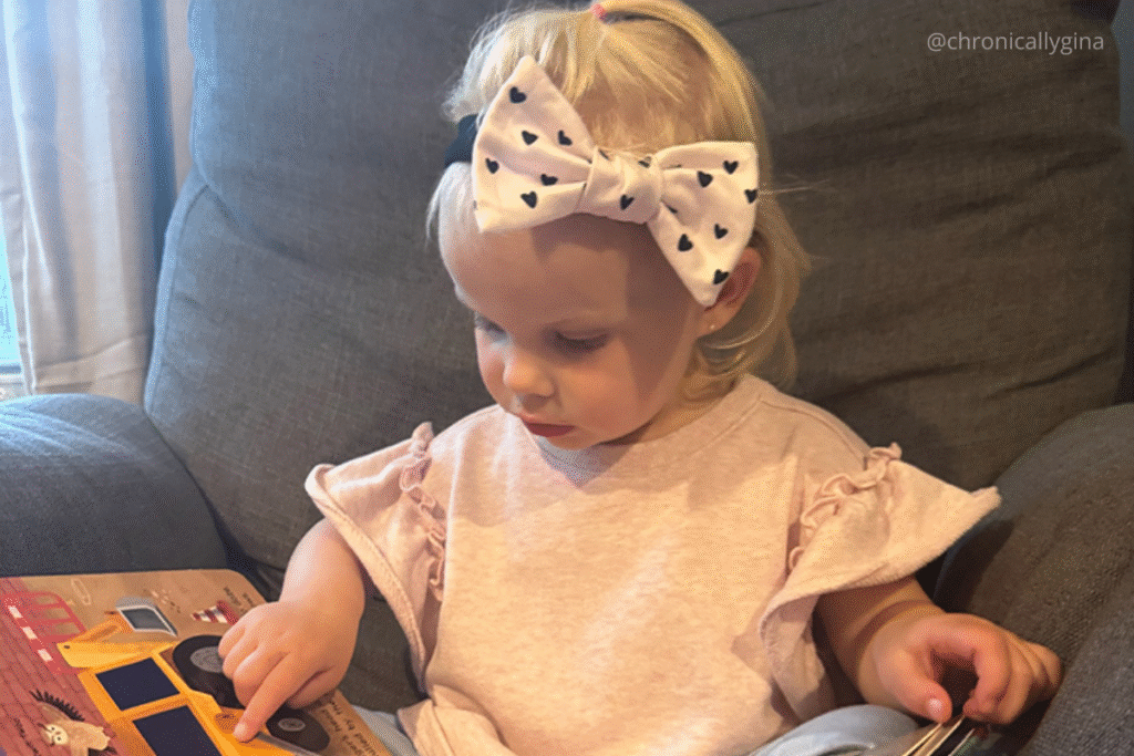 Toddler wearing a light pink shirt and a bow headband with black hearts, sitting on a gray chair and reading a children’s book—an adorable budget-friendly gift idea.