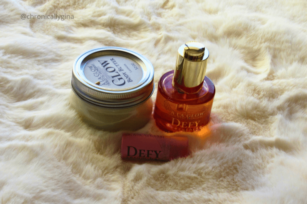 A’ La Glow Holistic Skincare products including a jar of Glow Body Butter, a bottle of Defy anti-aging serum, and a Defy lip balm displayed on a soft cream fur background.